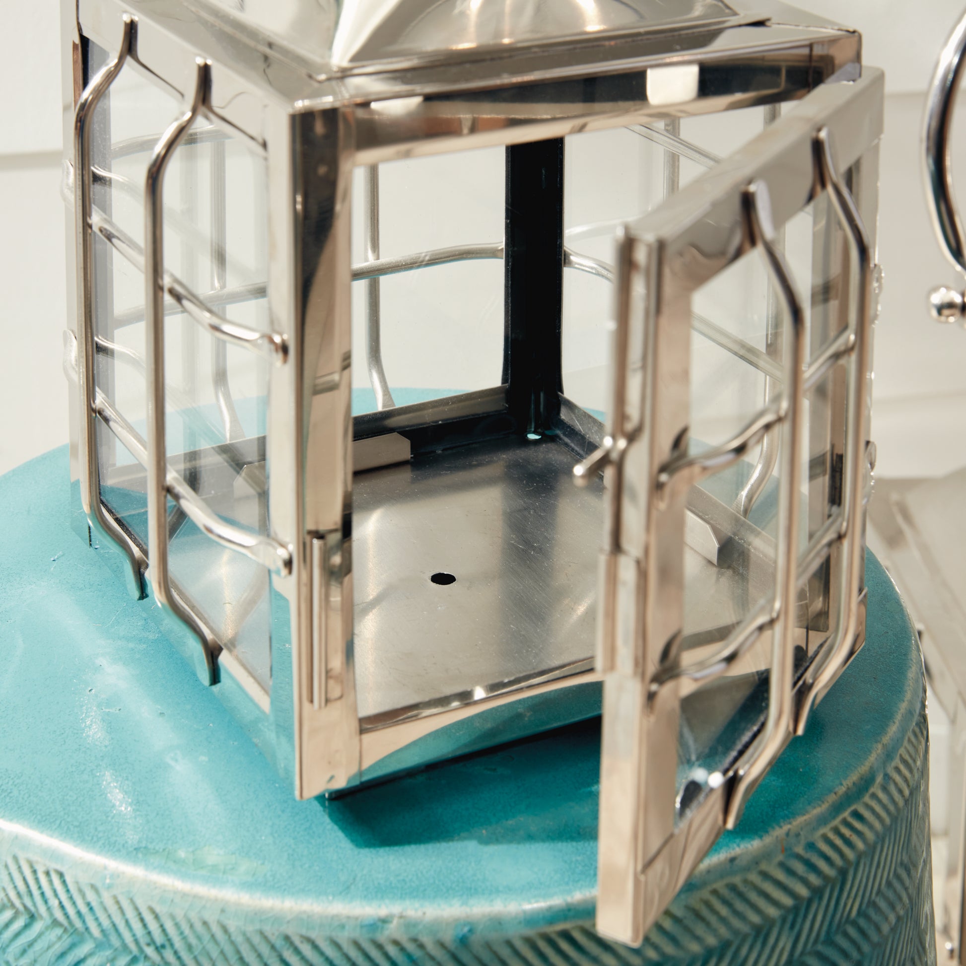 Metal lantern with glass panels and an open door, resting on a textured teal surface.