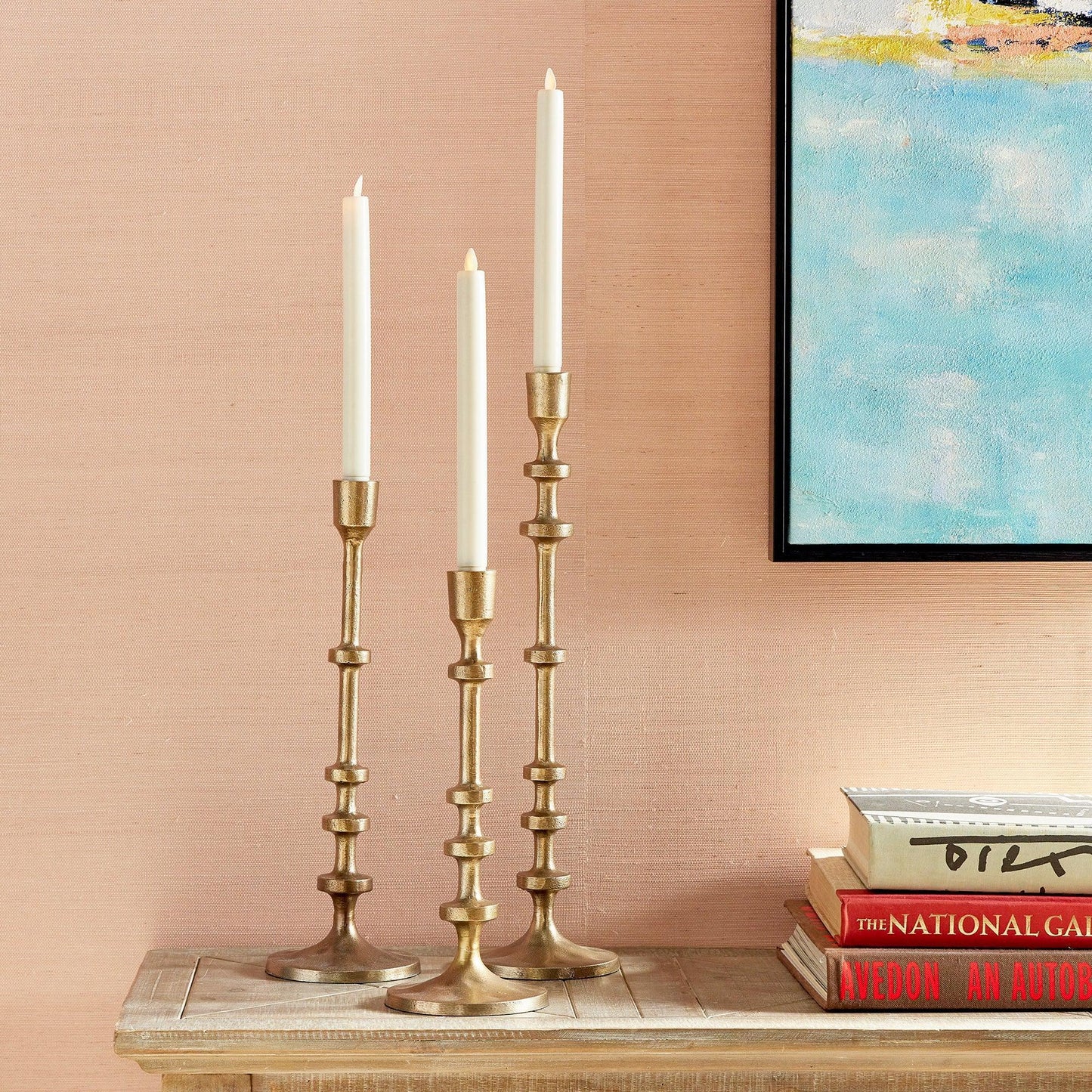 three gold taper candle holders with candles on wood table with books and a painting