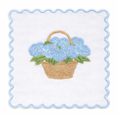 White cocktail napkin embroidered with blue hydrangeas in a tan basket, green leaves, and a light blue scalloped border