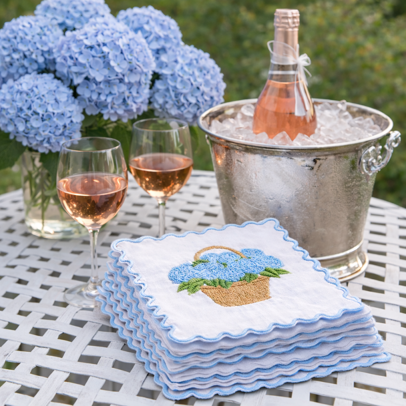 Stacked blue hydrangea embroidered cocktail napkins on an outdoor table beside rosé glasses, an ice bucket, and fresh hydrangeas