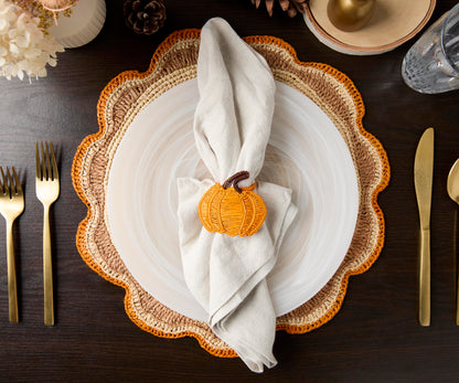 Table setting with a decorative plate, woven placemat, golden cutlery, and a pumpkin napkin holder.