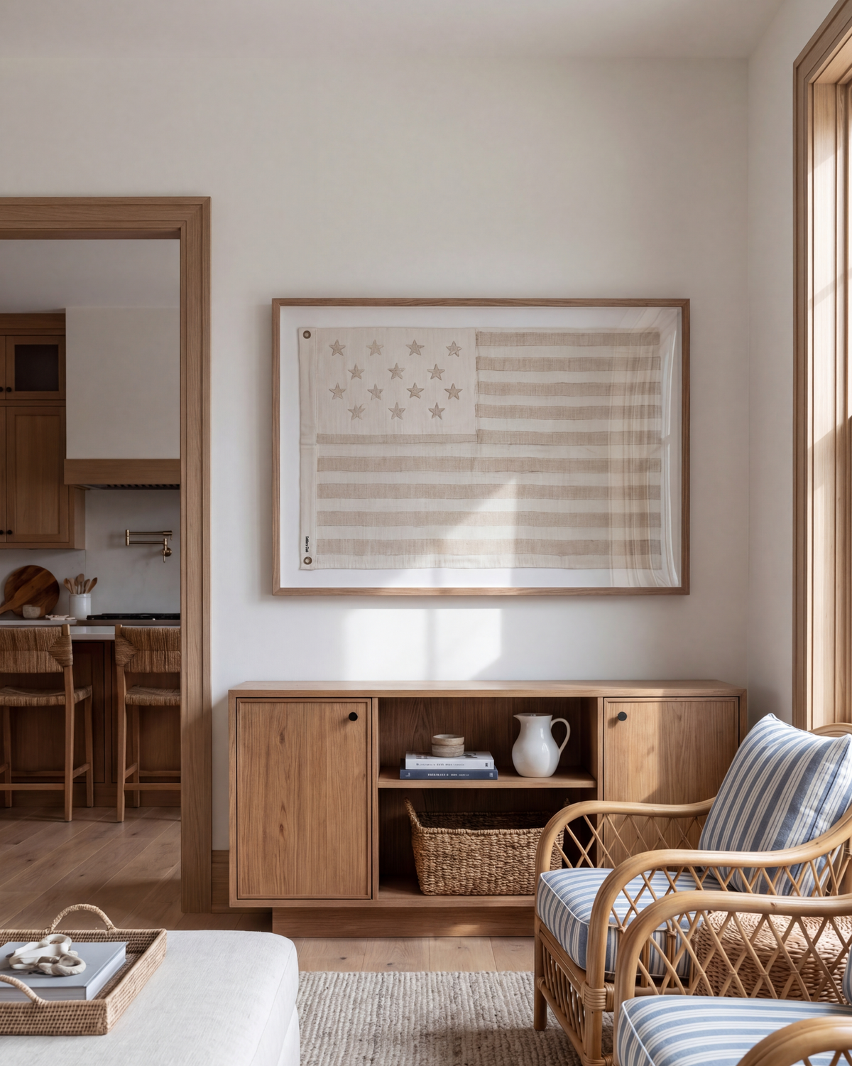Framed sand-toned American flag above a low wood cabinet beside a woven armchair.