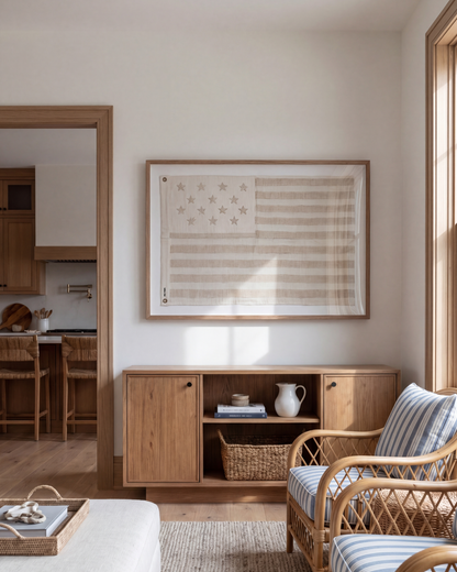 Framed sand-toned American flag above a low wood cabinet beside a woven armchair.