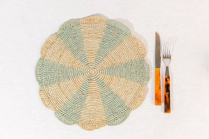 Round woven placemat in green and beige with a fork and knife beside it on a light surface.
