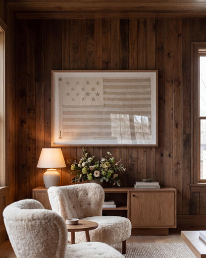 Framed sand-toned American flag above a cabinet in a wood-paneled room with cream armchairs.