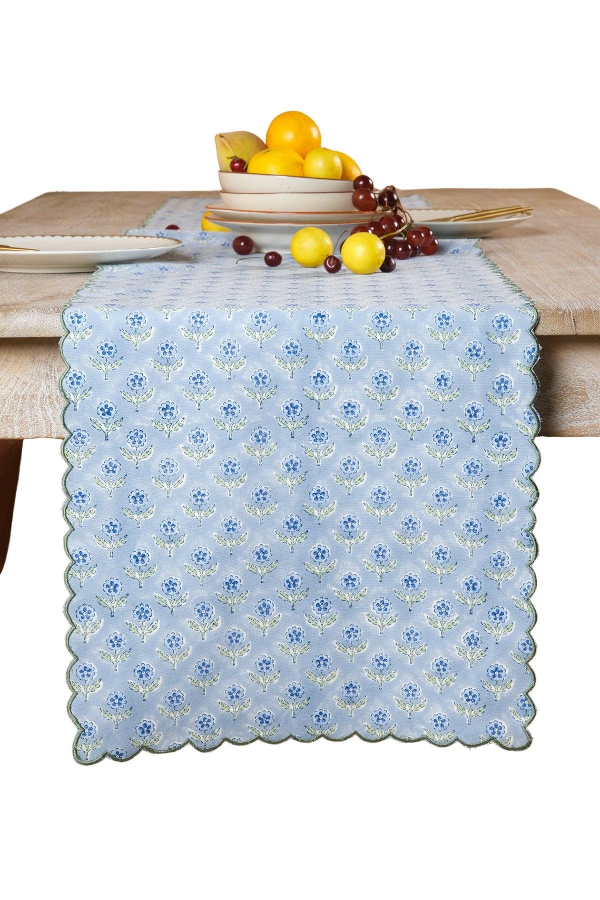 Mona pale blue floral table runner with green embroidered scalloped edges draped over a wood table, styled with dishes and fruit.