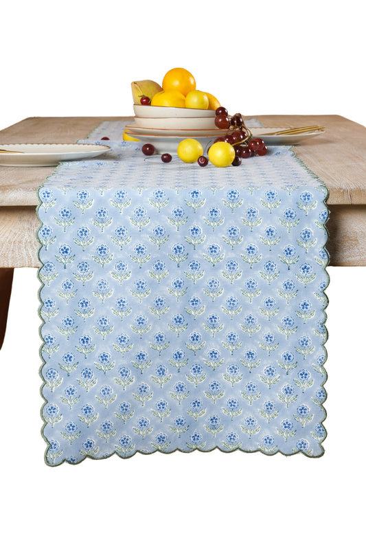 Mona pale blue floral table runner with green embroidered scalloped edges draped over a wood table, styled with dishes and fruit.