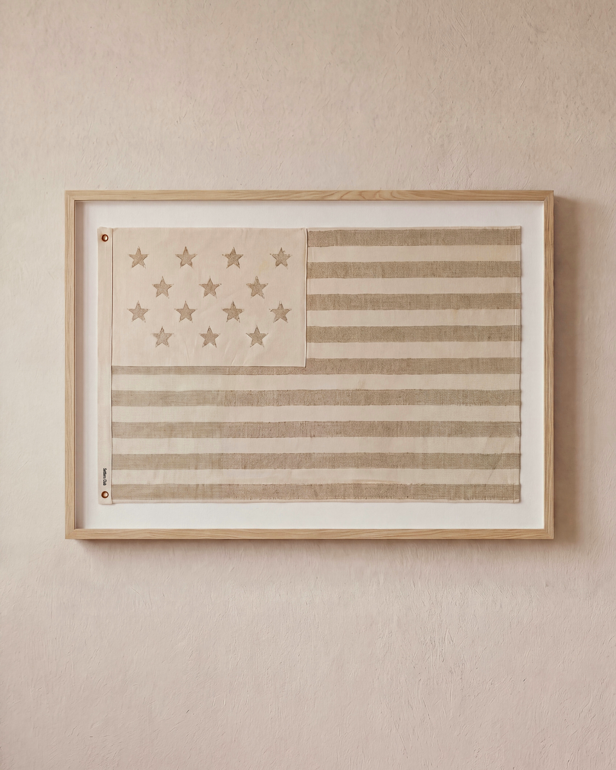 Sand-toned embroidered American flag displayed horizontally in a wood shadowbox frame on a neutral wall.