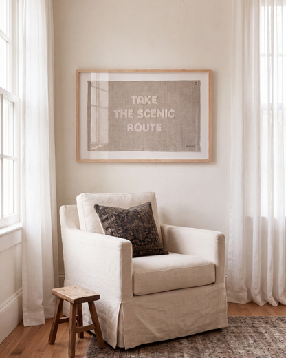 Scenic Route embroidered wall art hanging above a cream armchair in a bright reading nook