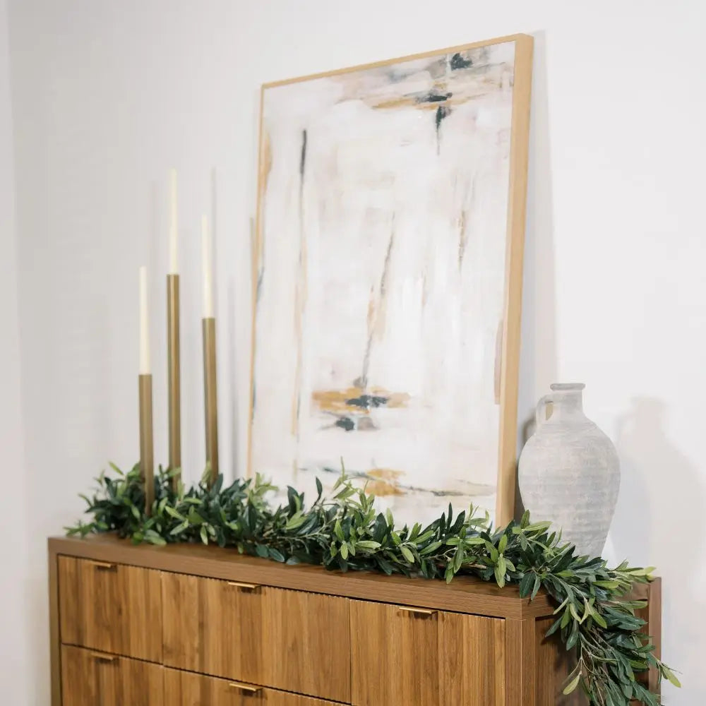 Gold-finish iron taper candle holders styled on a wooden console with greenery beneath abstract artwork
