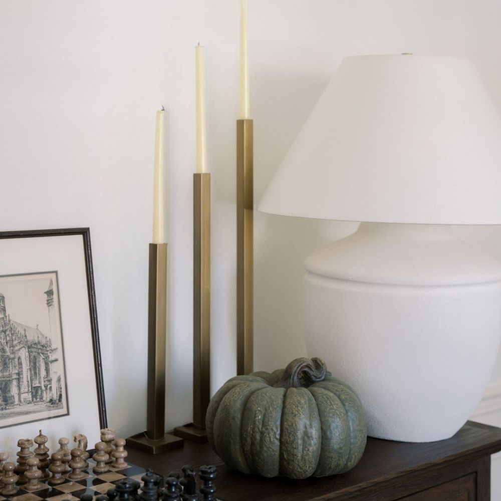 Three gold-finish iron taper candle holders styled with ivory candles beside a white lamp and green pumpkin