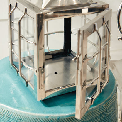 Metal lantern with glass panels and an open door, resting on a textured teal surface.