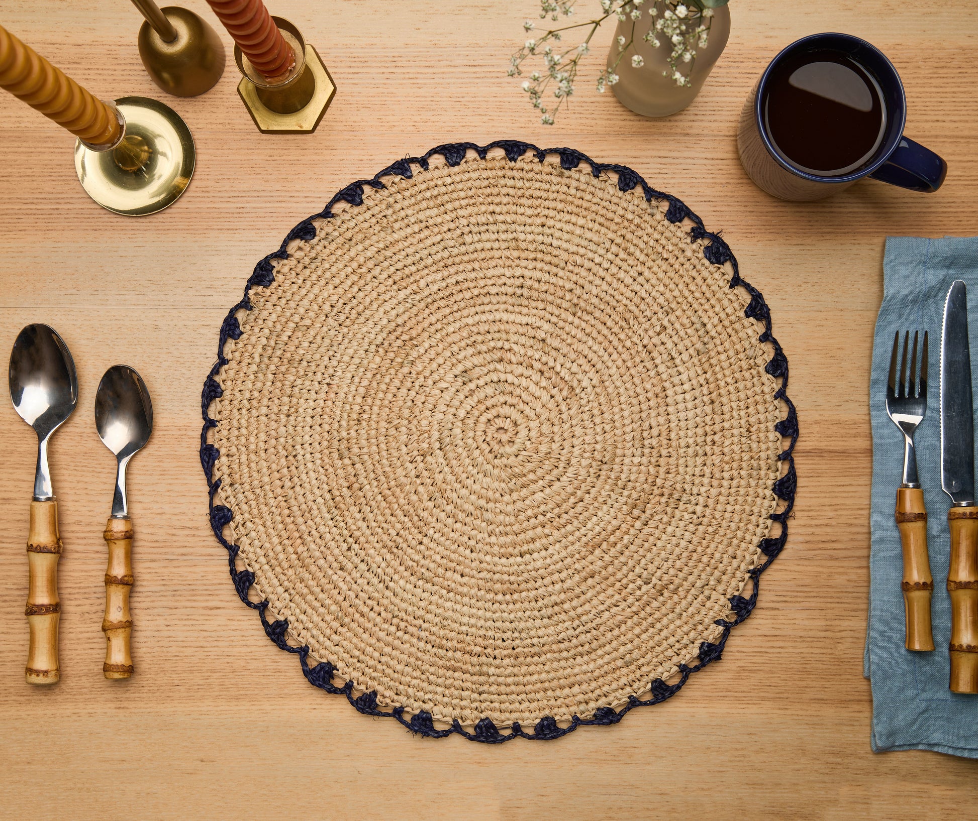 Round woven placemat on a wooden table with cutlery and a cup.