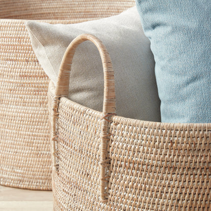 Woven wicker basket with a handle, holding soft, textured cushions in neutral tones.