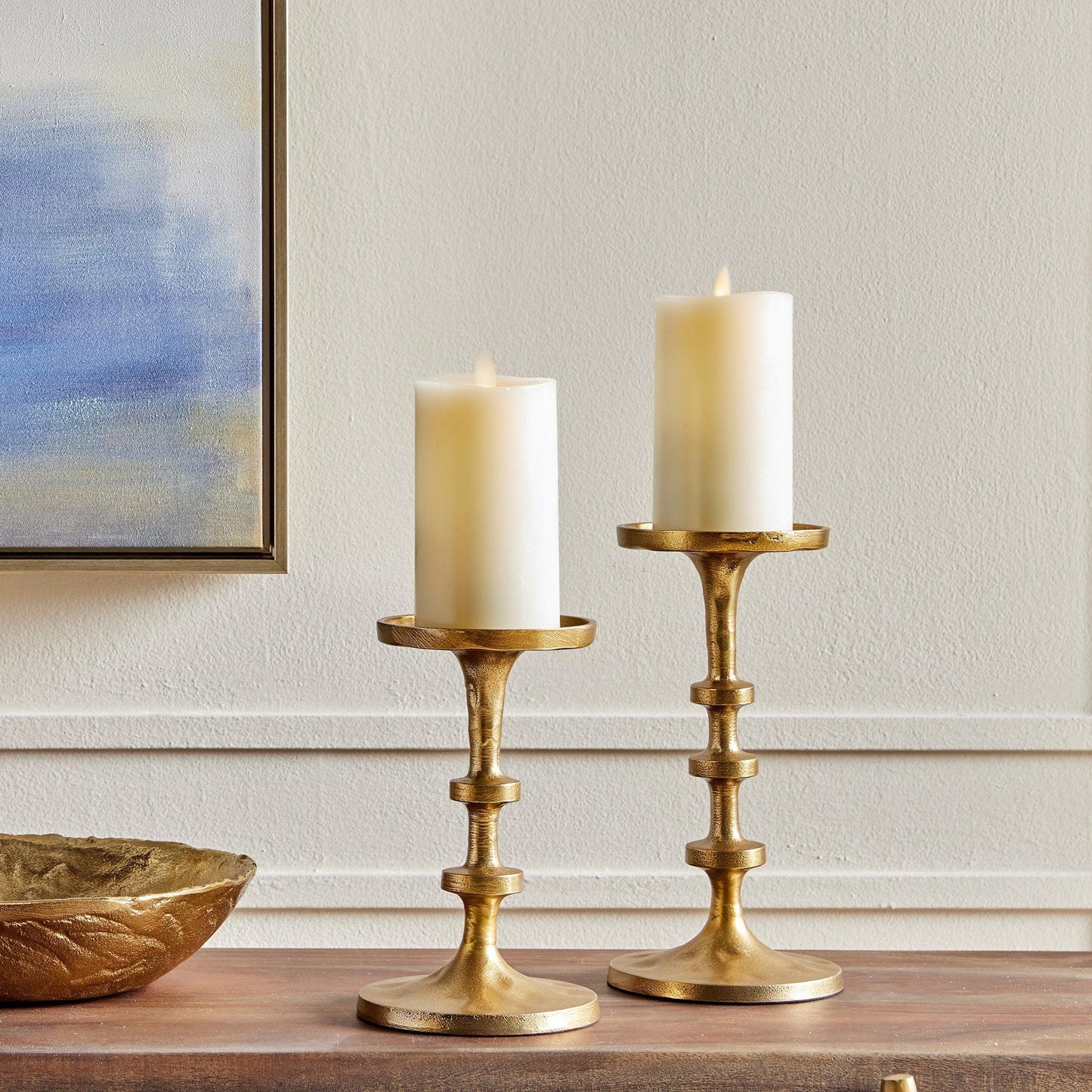 two gold candle stands with pillar candles on a table with a painting behind it