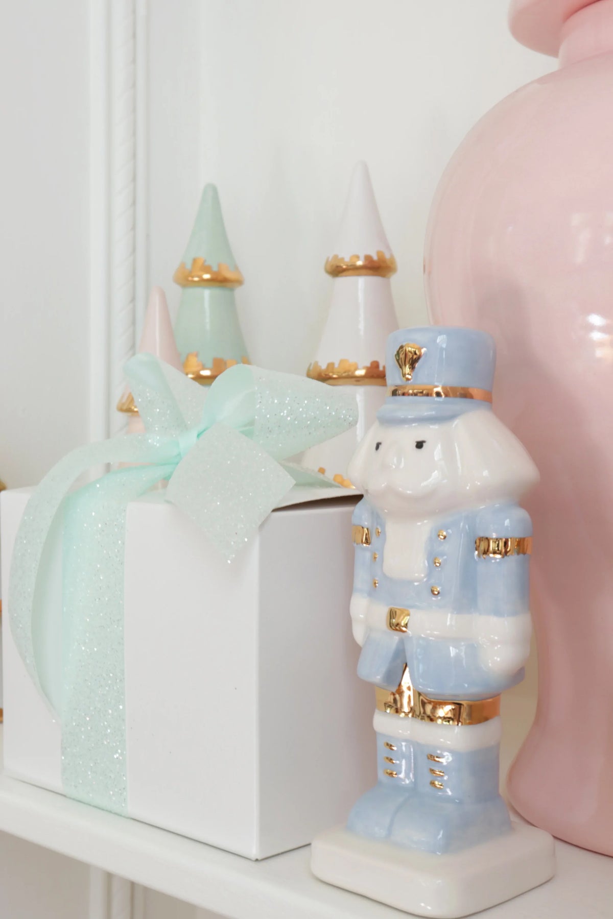 Light blue nutcracker styled beside a white gift box with a mint bow, pastel trees, and a pink jar