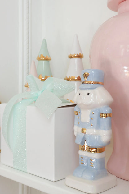 Light blue nutcracker styled beside a white gift box with a mint bow, pastel trees, and a pink jar