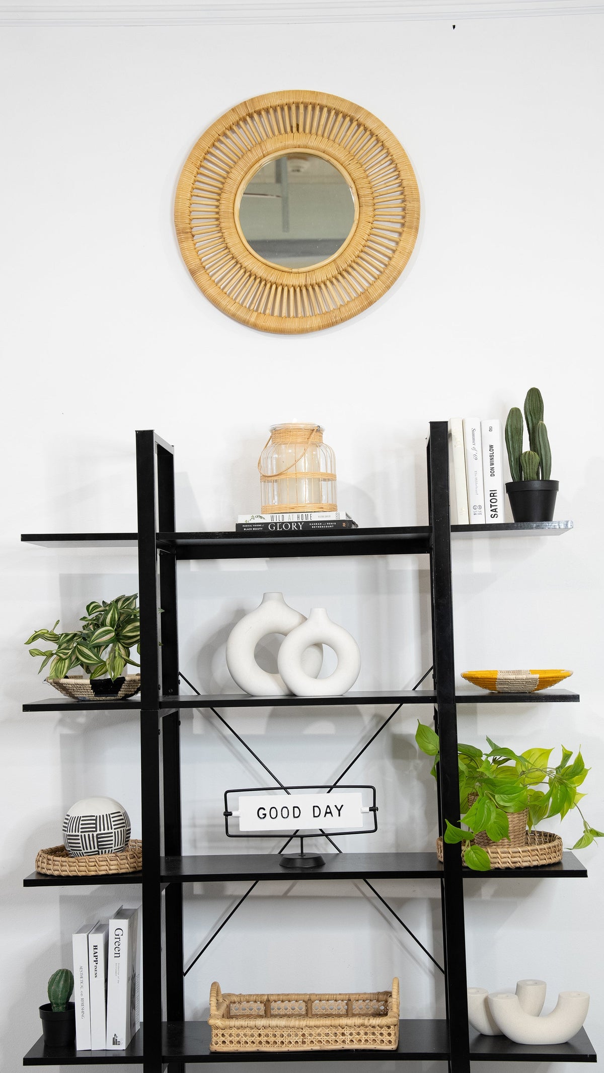 Natural round rattan wall mirror hanging above black shelving styled with books, plants, and decorative objects