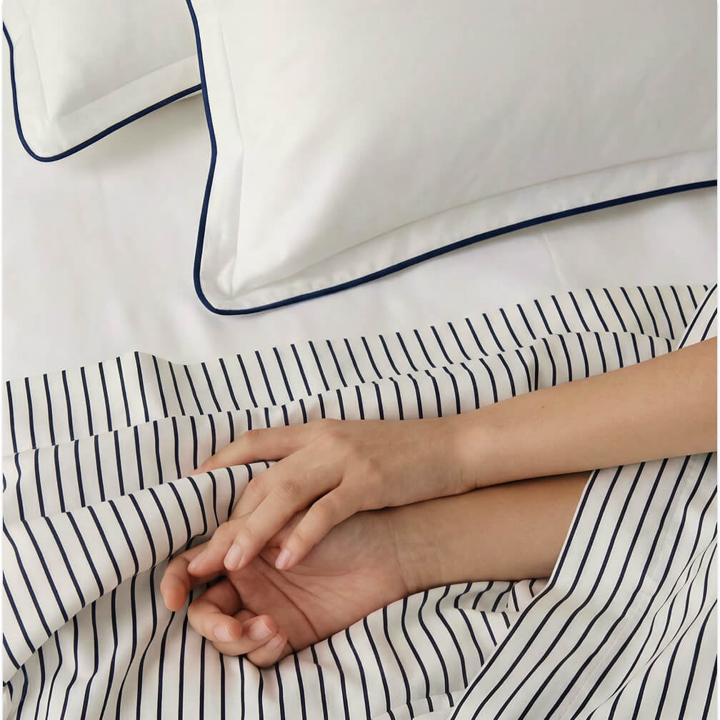 Porto blue-trimmed white pillowcases above striped bedding with a person's hands resting on the sheet